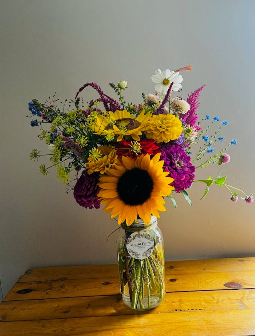 Floral arrangement in a glass jar on a wooden table featuring sunflower, zinnia, and other colorful flowers.