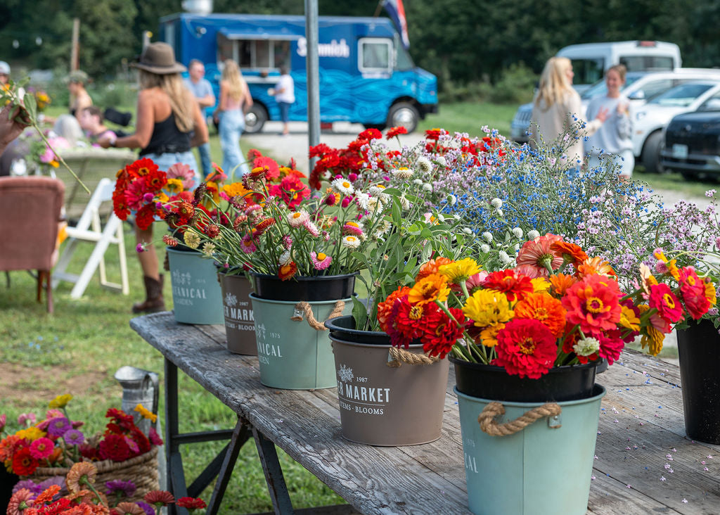 Buckets of brightly colored flowers outside on a work table with people assembling arrangements in the background.