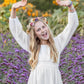 Happy girl in a white dress with her hands in the air posing for a photo in a field of purple flowers with a handmade dried-flower crown on her head.