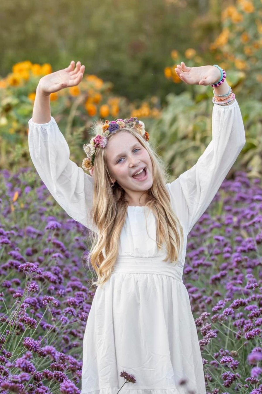 Happy girl in a white dress with her hands in the air posing for a photo in a field of purple flowers with a handmade dried-flower crown on her head.